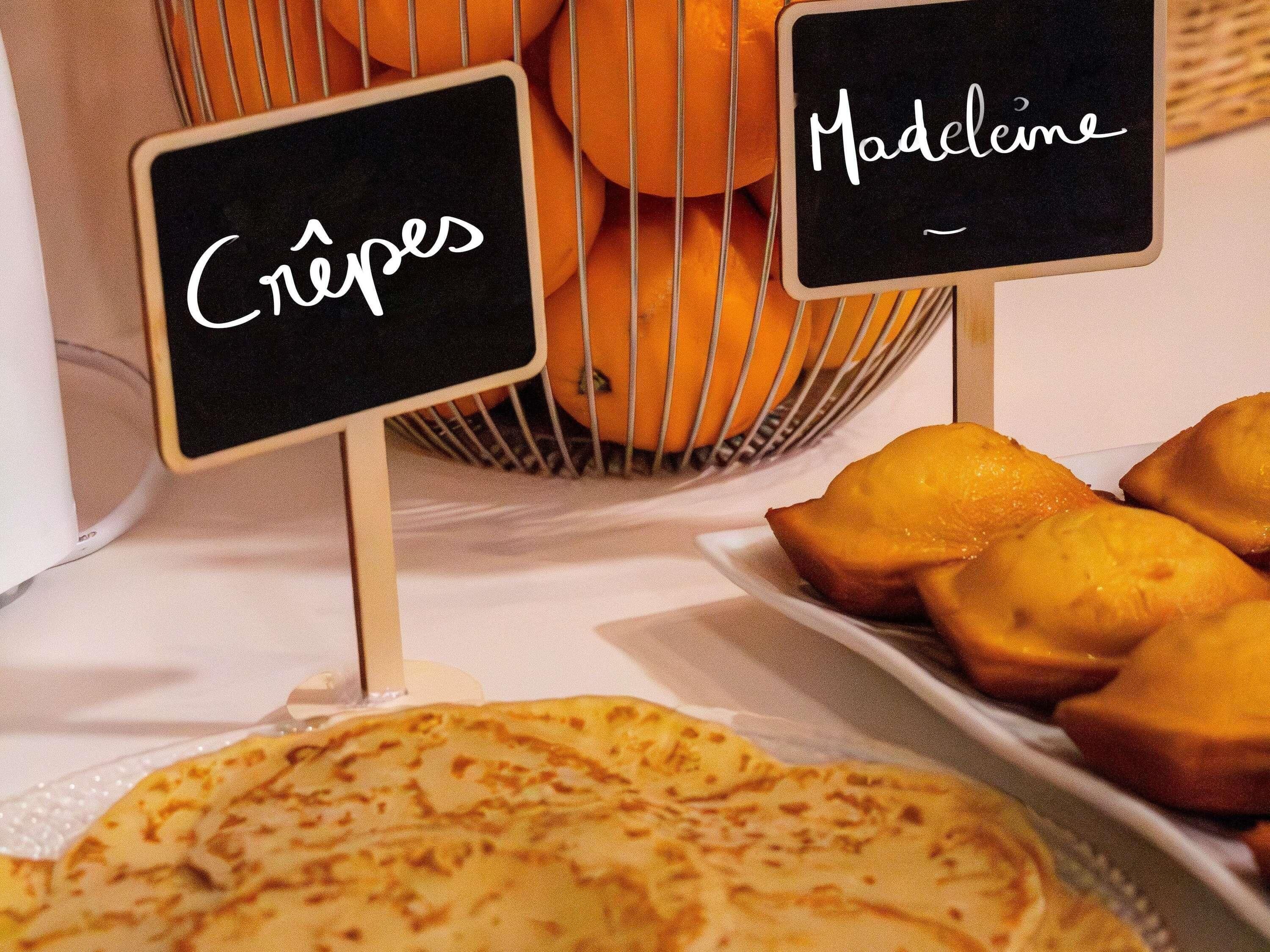 Eine Dessertauslage mit Schildern, auf denen 'Crêpes' und 'Madeleine' stehen, und ein Korb Orangen im Hintergrund.