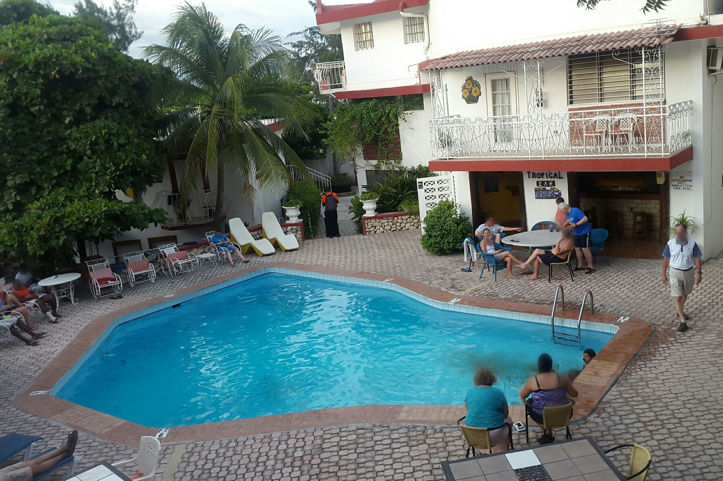 A swimming pool area with lounge chairs and tables, a Tropical Bar, and a building with a balcony.