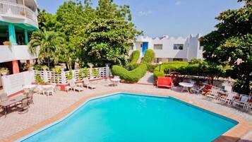 A swimming pool with white lounge chairs and tables, surrounded by greenery and a white fence.