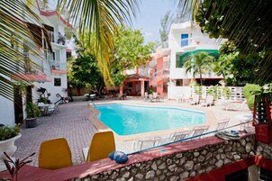 Una piscina rodeada de mobiliario exterior y follaje, con edificios al fondo.