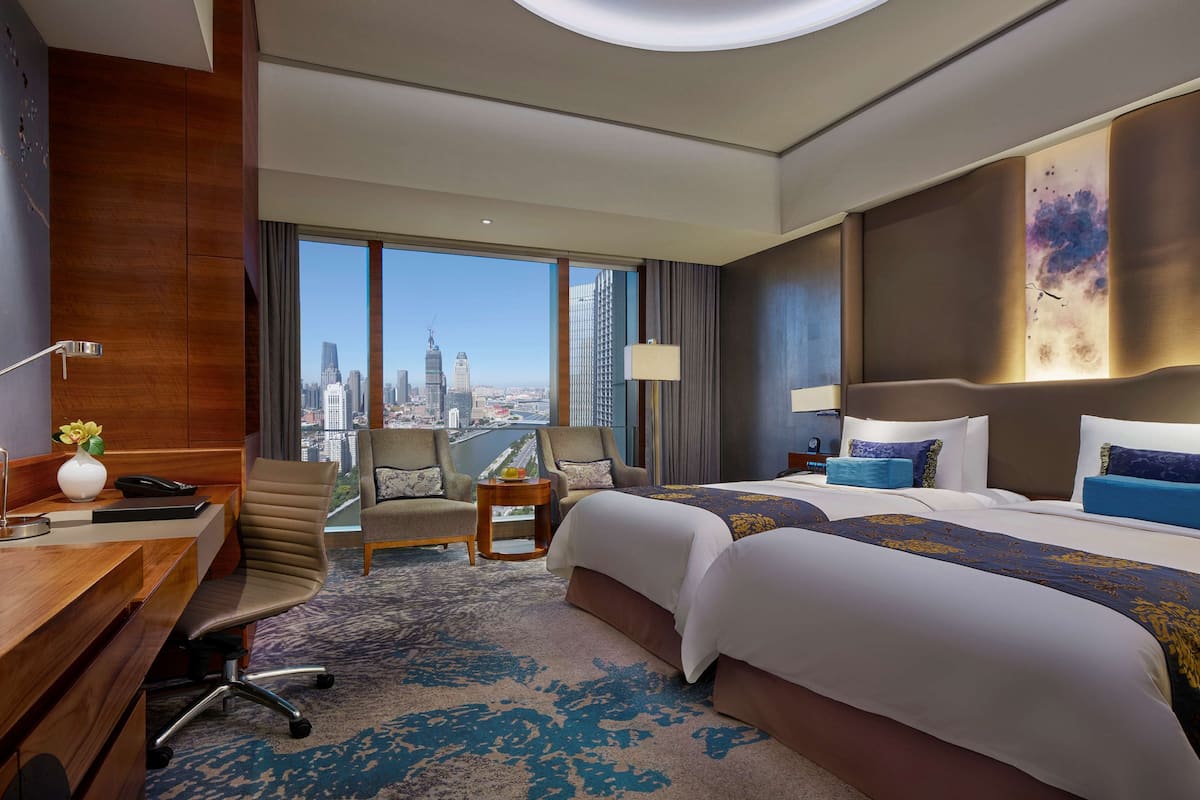 Quarto de hotel com duas camas, uma mesa, uma cadeira e vista para a cidade, além de uma janela grande.
