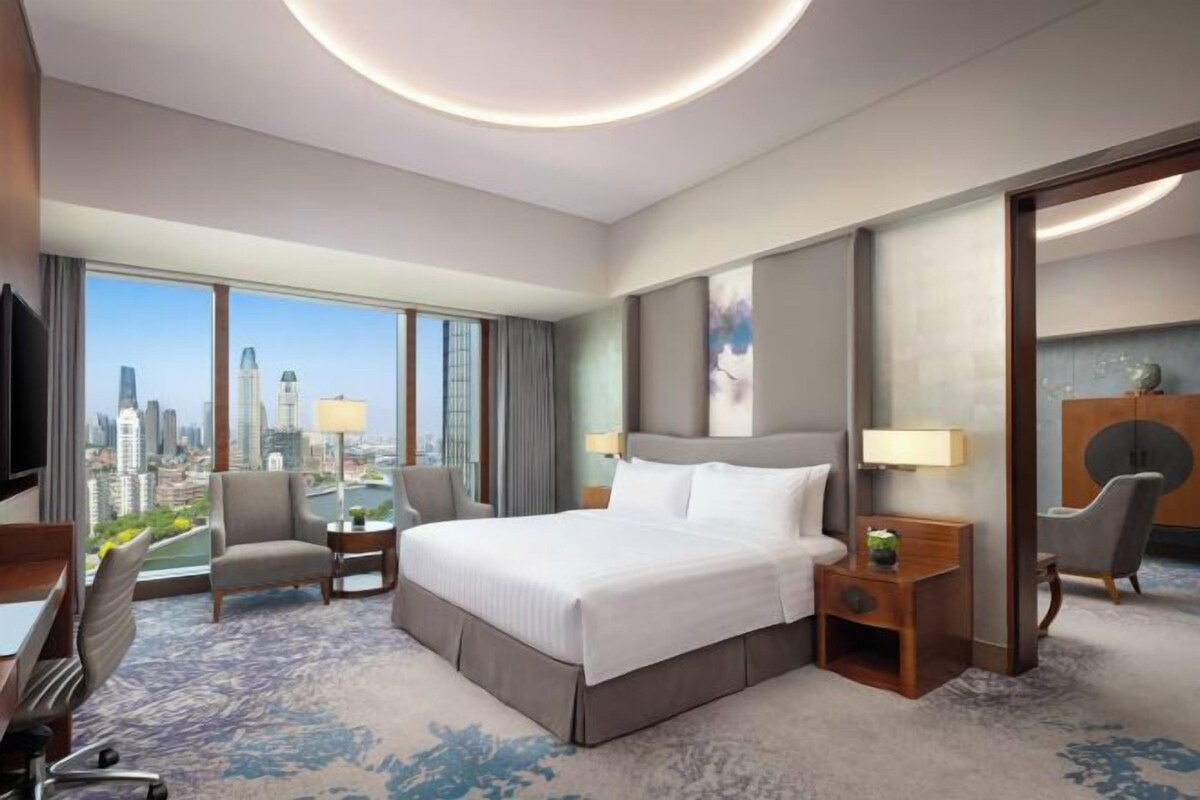 A hotel room with a large bed, a desk, a chair, a TV, and a view of the cityscape.