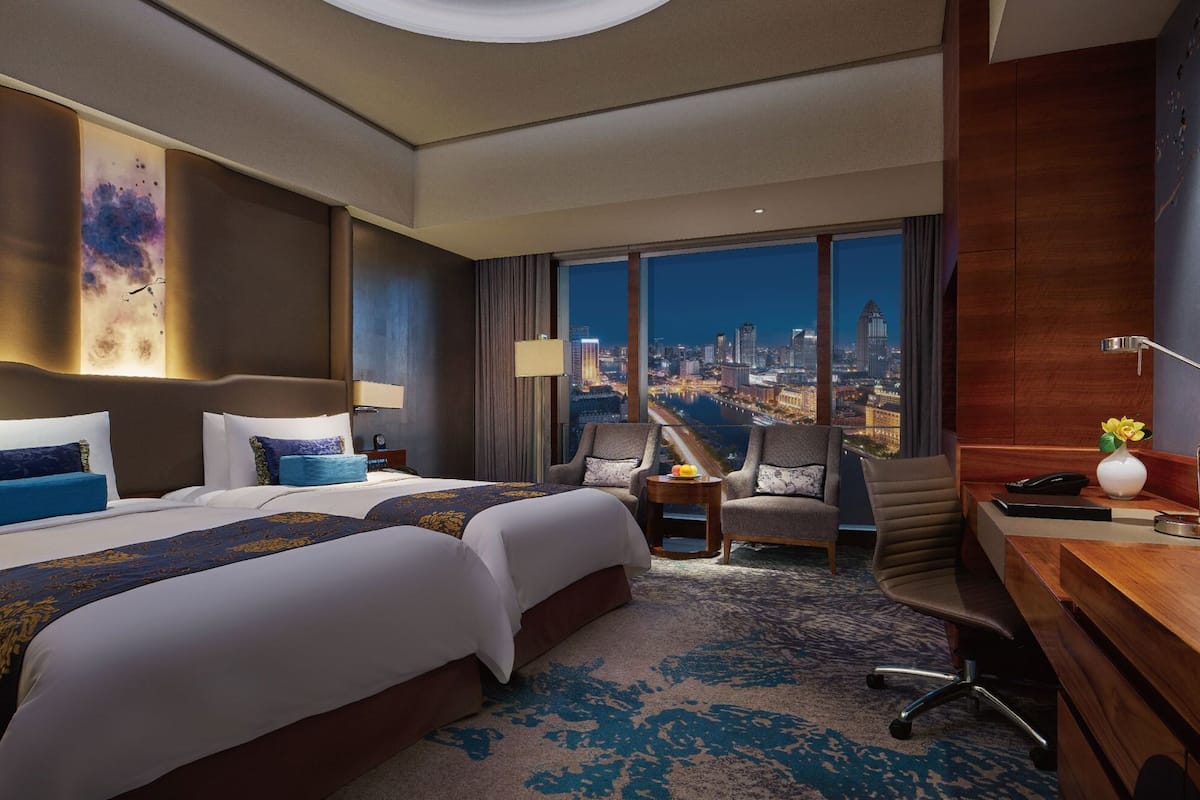 Quarto de hotel com uma cama grande, duas mesas de cabeceira, uma escrivaninha, uma cadeira e vista para a paisagem urbana.