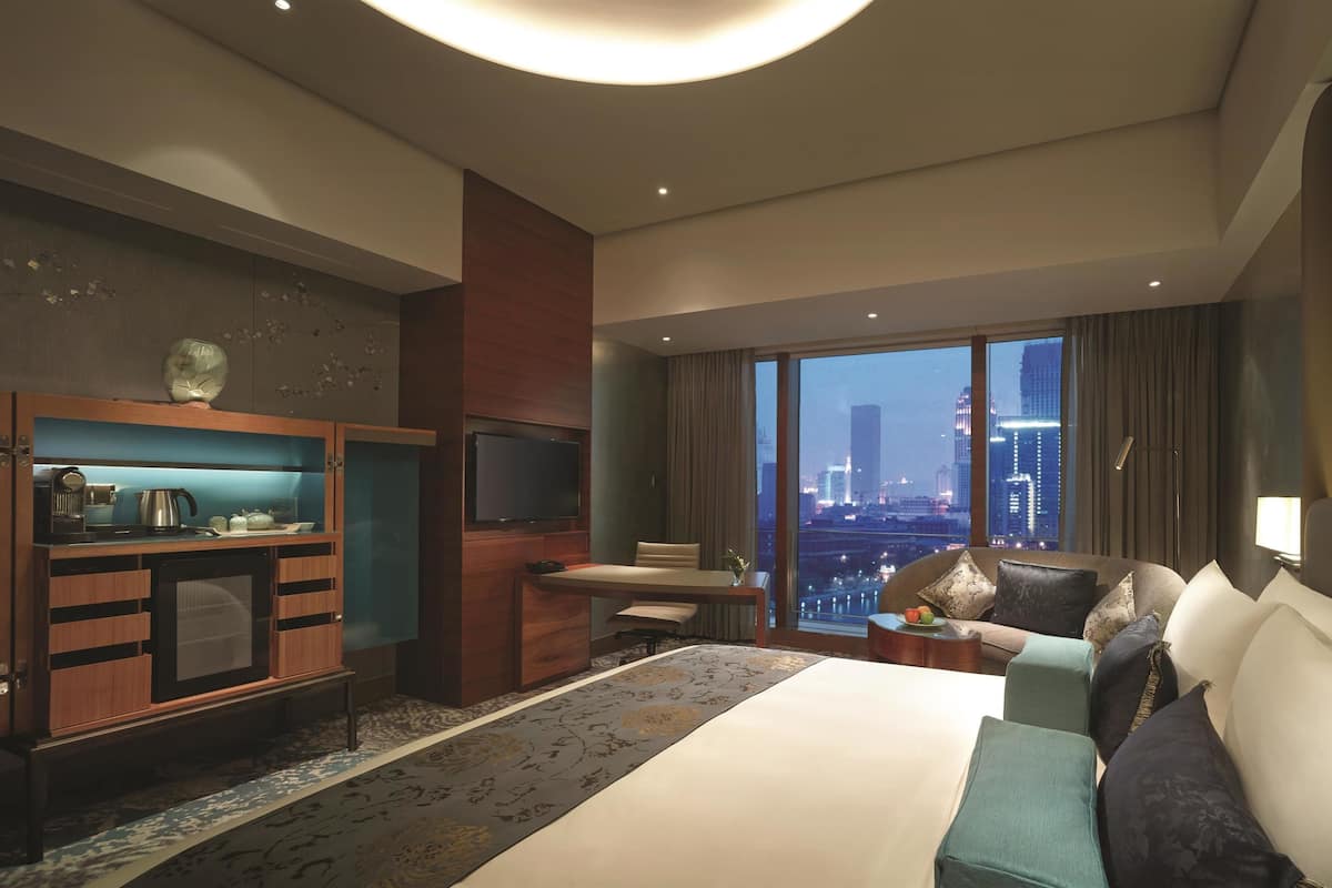 Quarto de hotel com uma cama grande, uma escrivaninha, televisão e vista para a paisagem urbana.