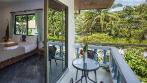 A hotel room with a bed, a balcony with a table and chairs, and a view of lush greenery. - Ya Nui Resort (Rawai)