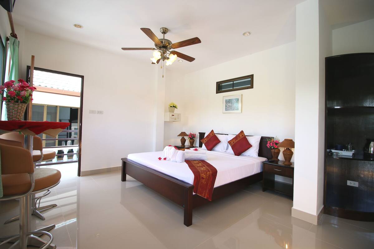 A hotel room with a large bed, a ceiling fan, a nightstand with a lamp, and a view of the outside through a window.
