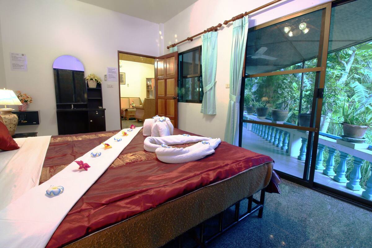 A hotel room with a bed, a window with a view of greenery, and a door leading to another room.