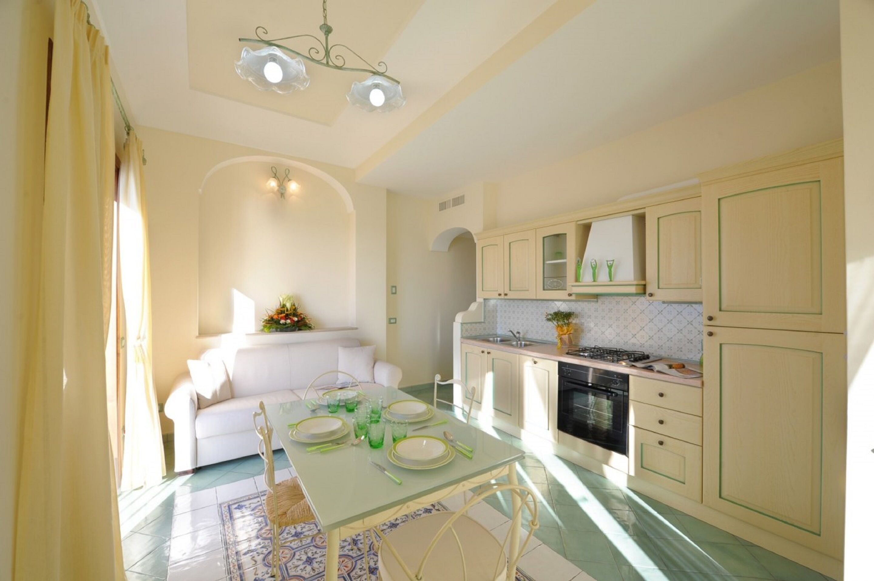 Una cocina compacta con zona de comedor, que presenta una mesa blanca para cuatro personas, un sofá al fondo y muebles de cocina en tonos claros.