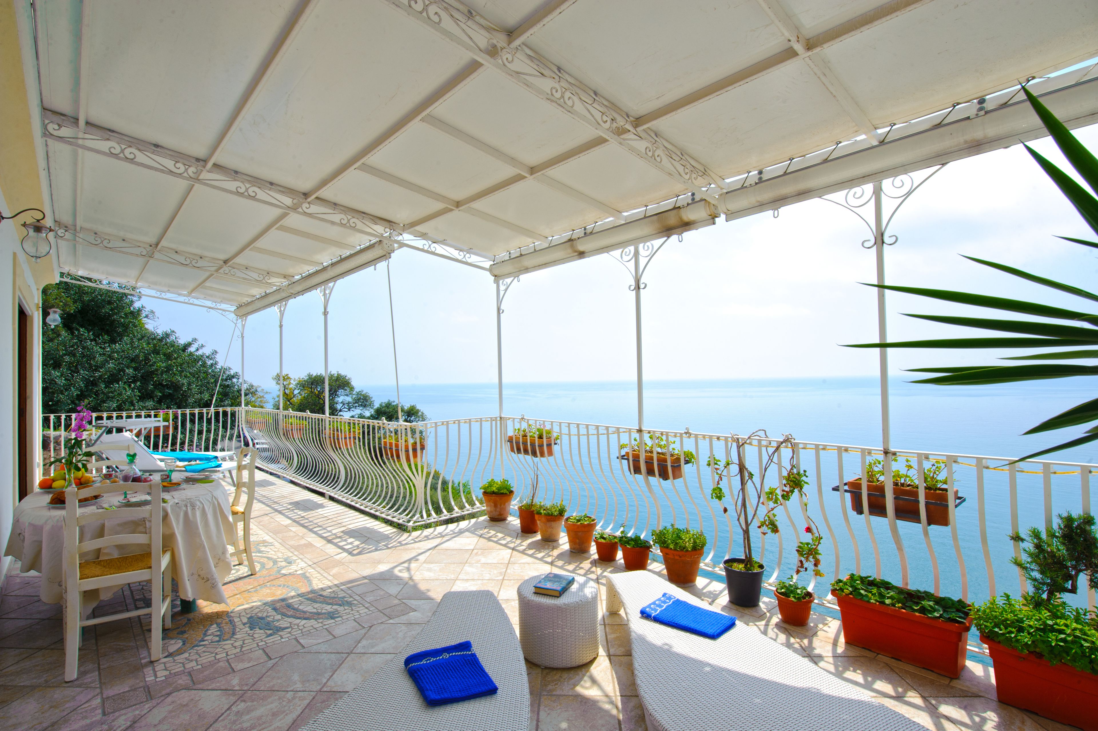Una terrazza coperta con vista sul mare, con un tavolo e sedie, piante in vaso e poltrone relax.