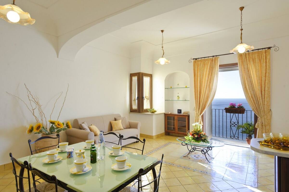 A dining area with a table set for four, a sofa, a cabinet, and a balcony with curtains.