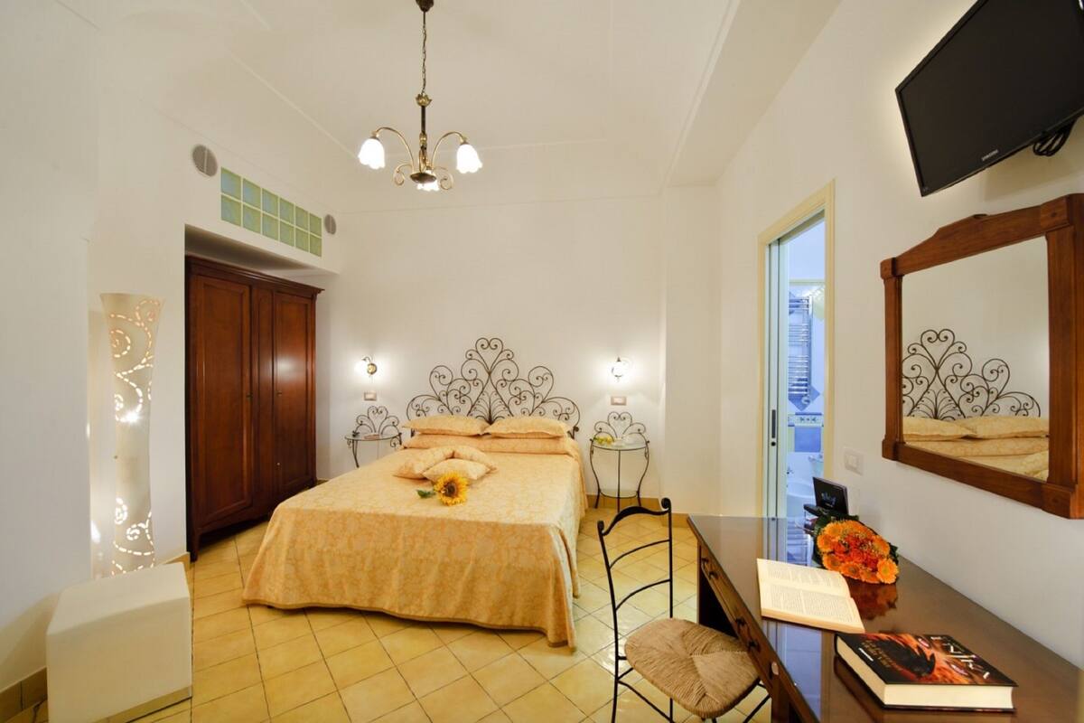 A bedroom with a bed, a desk with a chair, a TV, and a window.