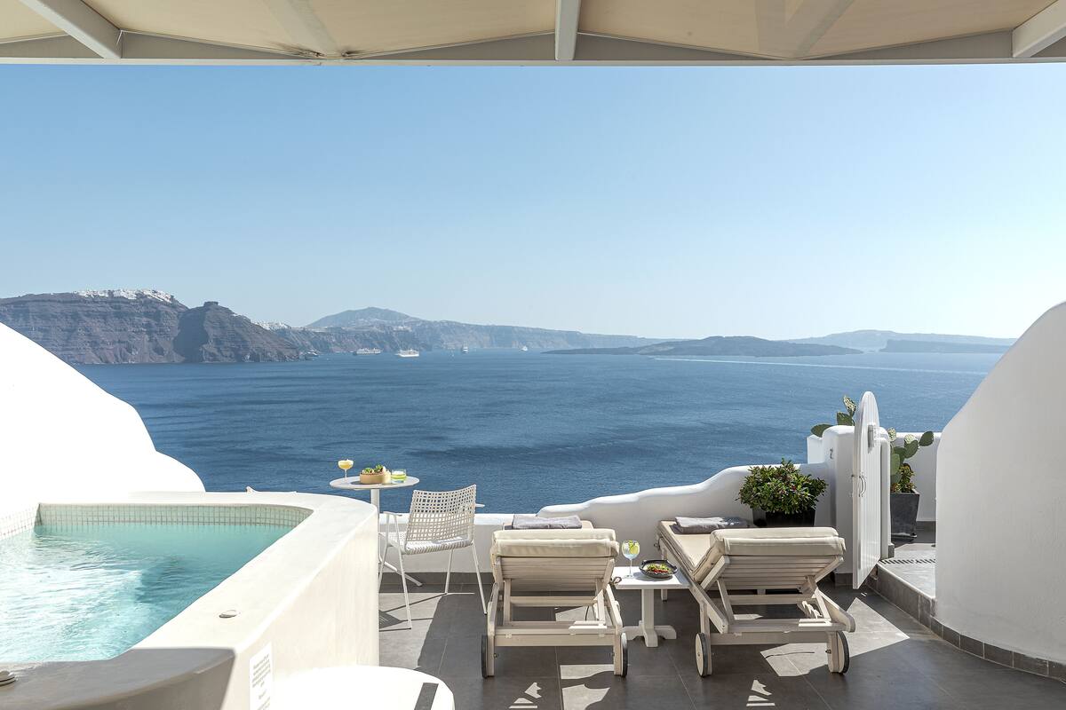 A sunlit terrace with a pool, lounge chairs, and a table with a drink, overlooking a scenic coastline.