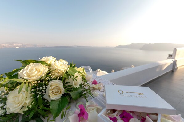 A table with a bouquet of white roses and a box labeled "SECRET HOTELS" on a terrace overlooking a body of water. - Santorini Secret Suites & Spa (Santorini)
