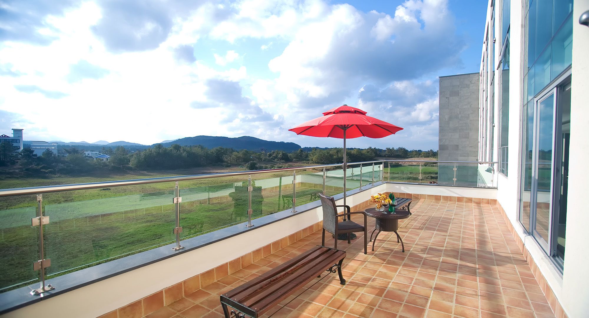 A balcony with a view of a golf course, a red umbrella, and a bench.
