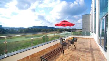 A balcony with a view of a golf course, a red umbrella, and a bench.