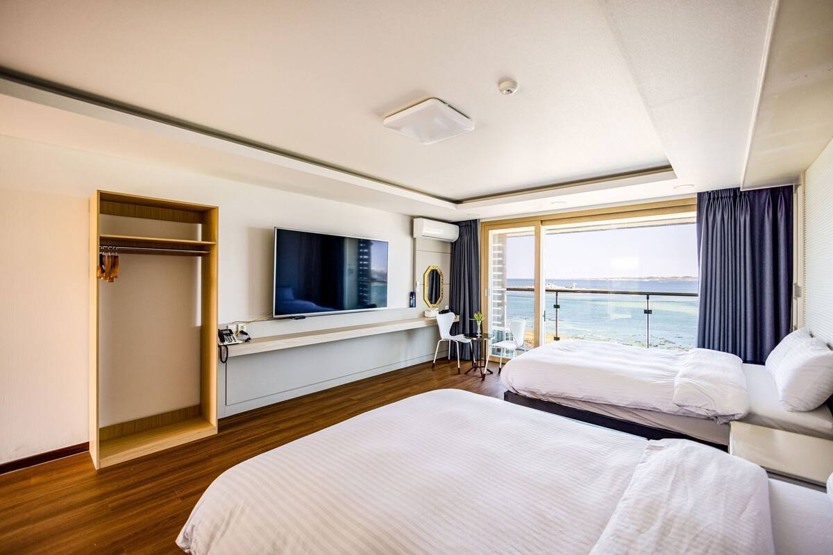 A hotel room with a large bed, a smaller bed, a TV, a desk with a chair, a wardrobe, and a balcony with a view of the sea.