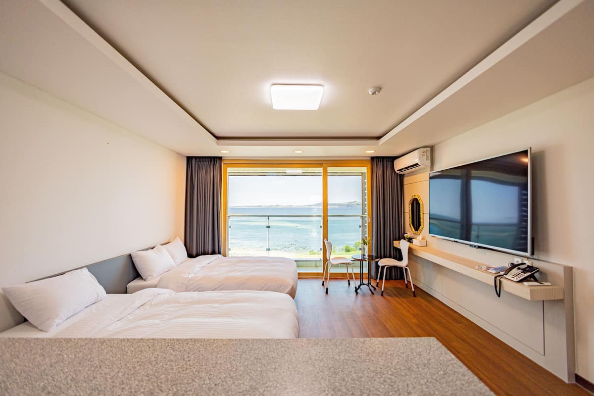 A hotel room with two beds, a TV, a desk with a chair, and a view of the sea.