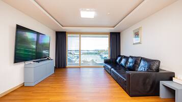 A modern living room with a large flat-screen TV, a leather sofa, and a sliding glass door leading to a parking area.
