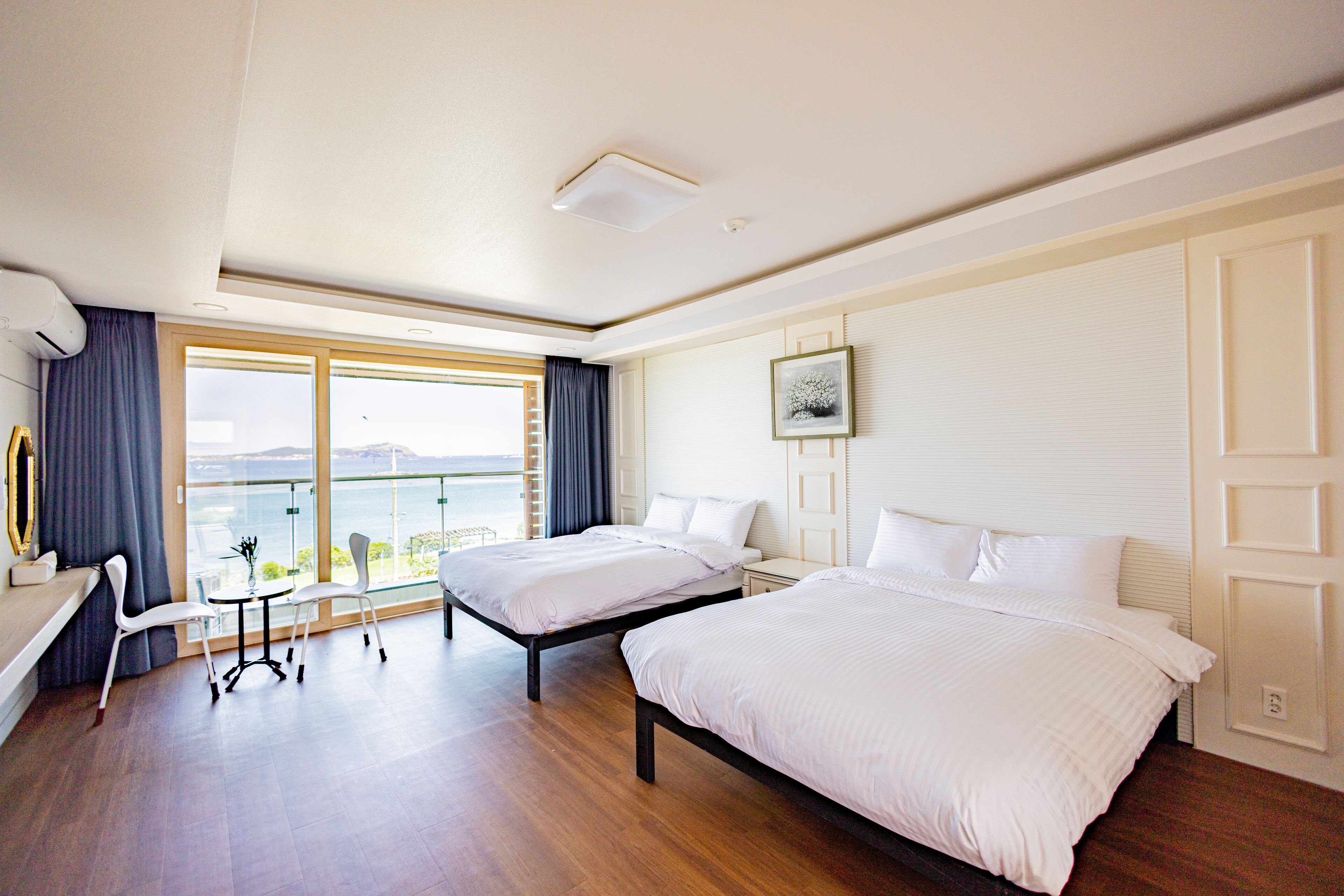 Habitación de hotel con dos camas, un escritorio y sillas, un ventanal con vistas y un cuadro en la pared.