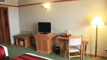 A hotel room with a bed, a desk with a chair, a television, a lamp, and a trash can.