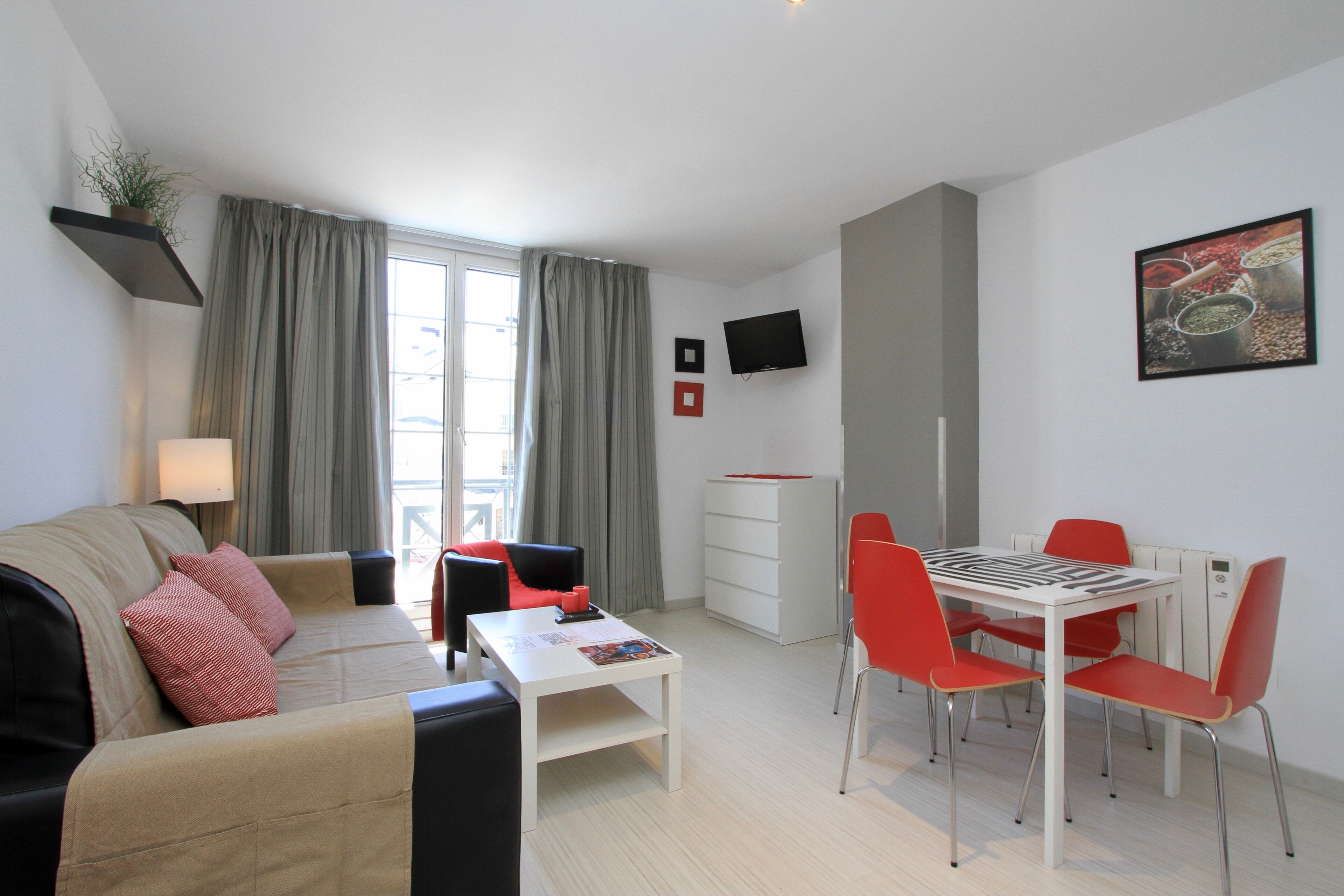 Ein modernes Wohnzimmer mit einer Couch, einem Esstisch mit Stühlen, einem an der Wand montierten Fernseher und einem Fenster mit Vorhängen.