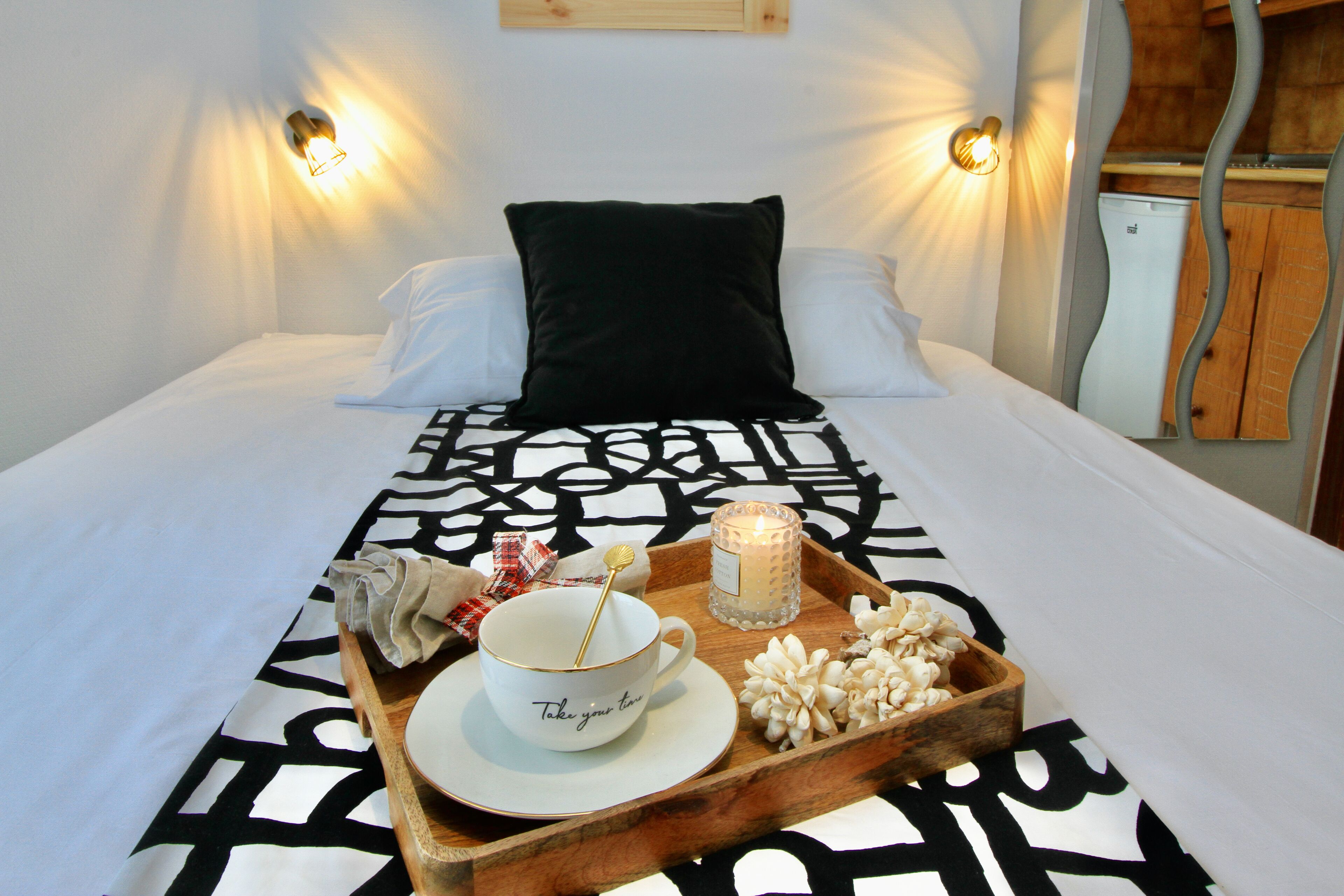 Un lit bien fait, avec un oreiller noir, un plateau contenant une tasse, une bougie et des fleurs décoratives posés sur le couvre-lit.