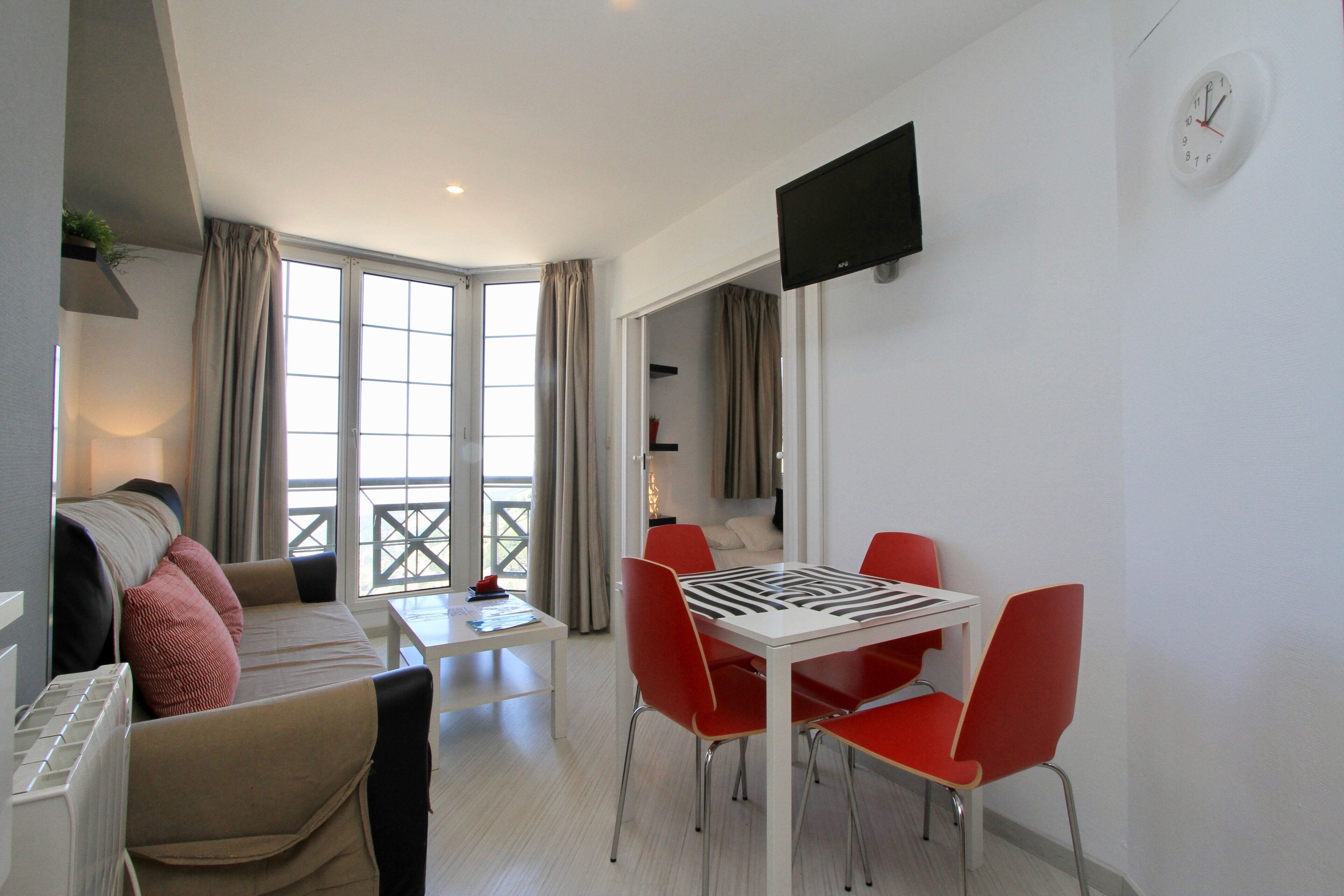 A modern living room with a sofa, a dining table with red chairs, a flat-screen TV, and a large window with curtains.