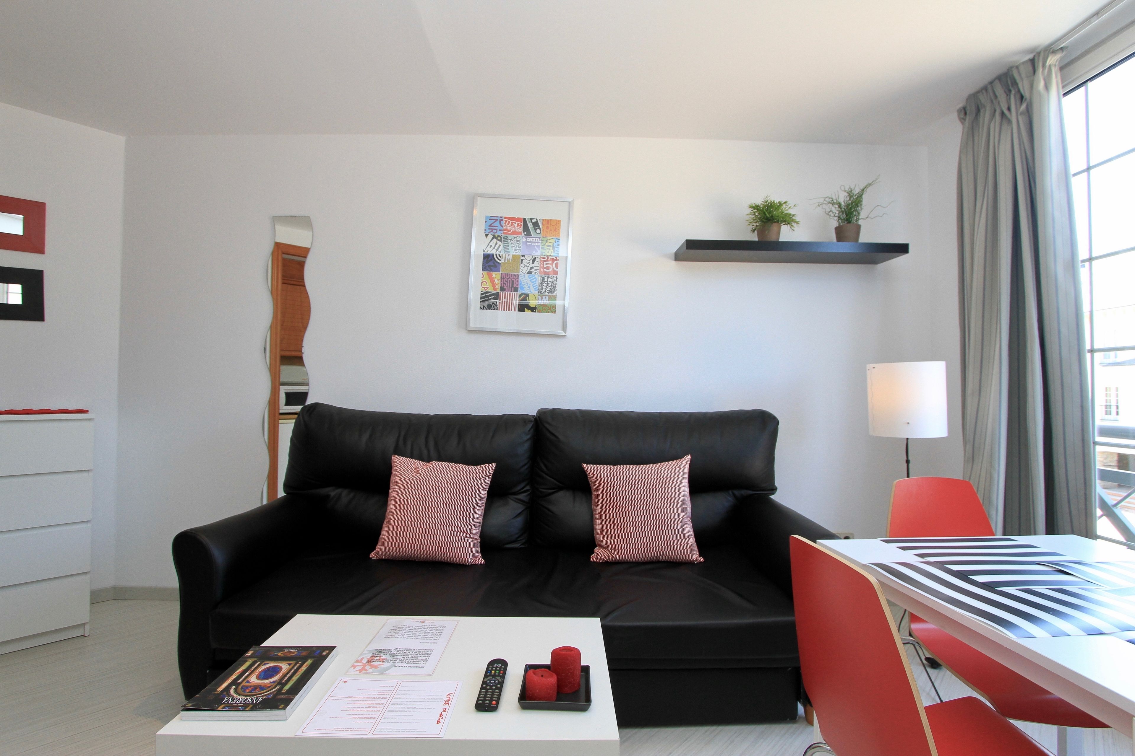 Een moderne woonkamer met een zwarte leren bank, twee rode kussens, een witte salontafel met een afstandsbediening en een boek, een rode fauteuil, een wit bureau met een toetsenbord, een lijst met een foto aan de muur en een raam met gordijnen.