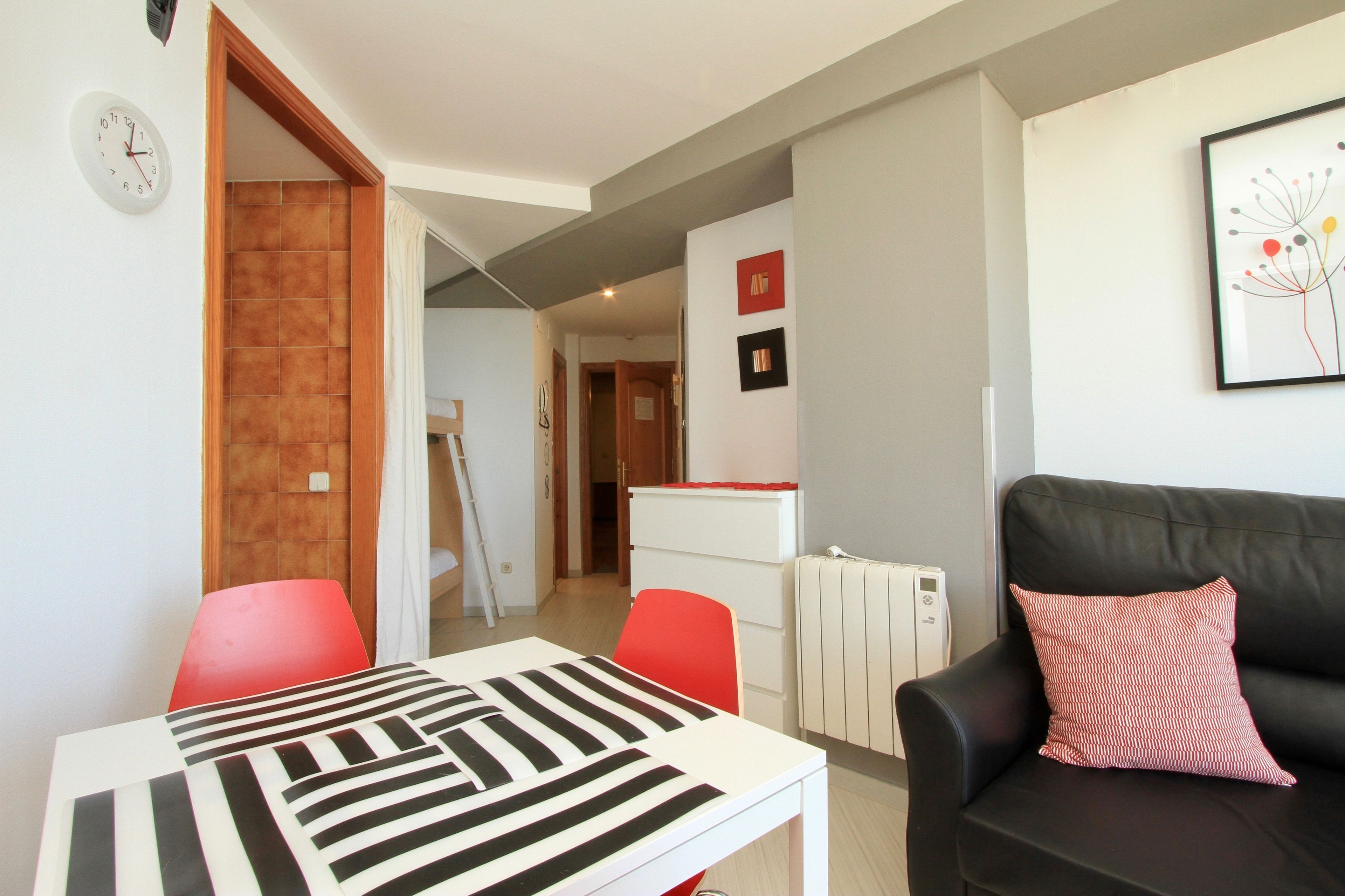 A modern living room with a dining table, red chairs, a black leather sofa, and a kitchen in the background.