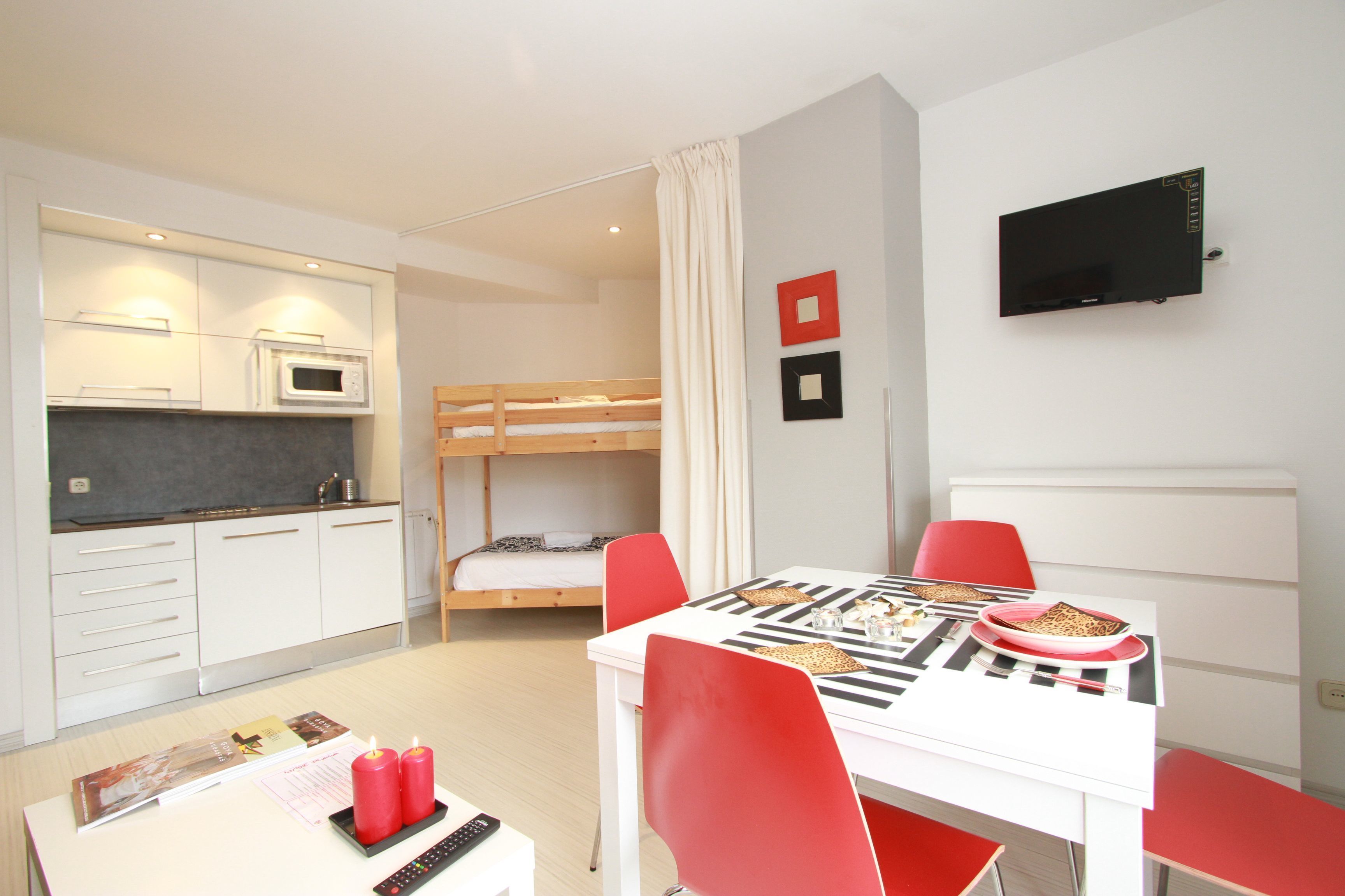A compact hotel room with a kitchenette, a single bed, a dining table with red chairs, and a flat-screen TV mounted on the wall.
