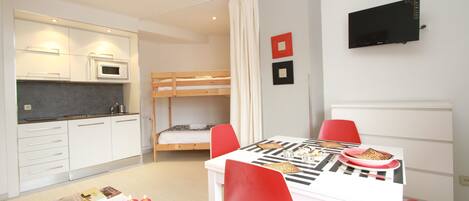 A compact hotel room with a kitchenette, a single bed, a dining table with red chairs, and a flat-screen TV mounted on the wall.