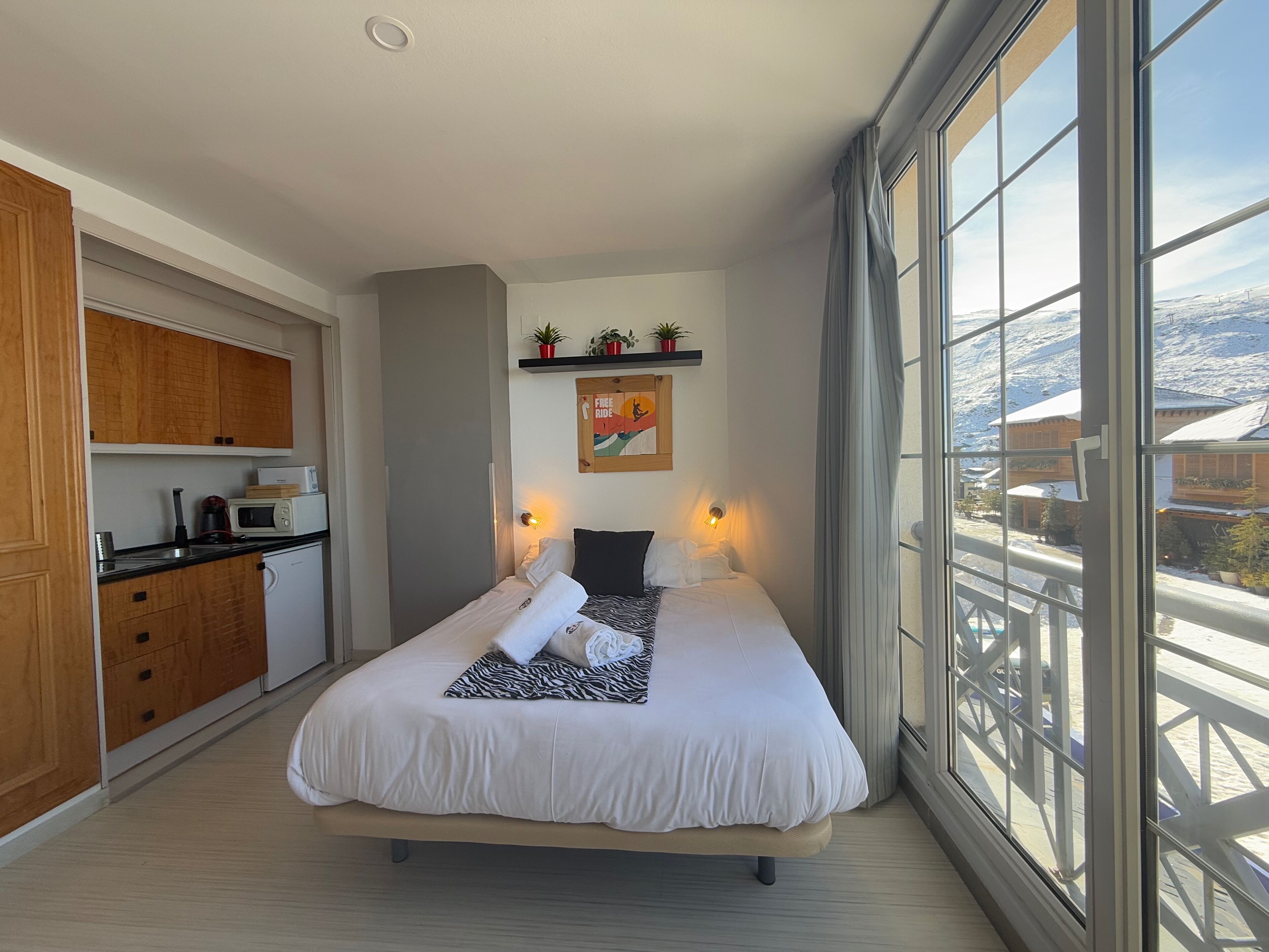 A bedroom with a bed, bedside lamps, a view of a snowy landscape, a kitchen area with cabinets and appliances, and a mounted picture on the wall.