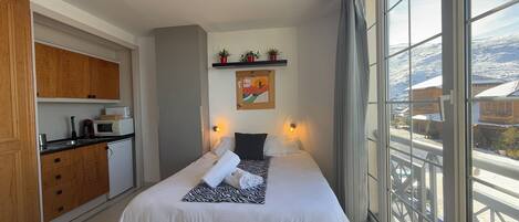A bedroom with a bed, bedside lamps, a view of a snowy landscape, a kitchen area with cabinets and appliances, and a mounted picture on the wall.