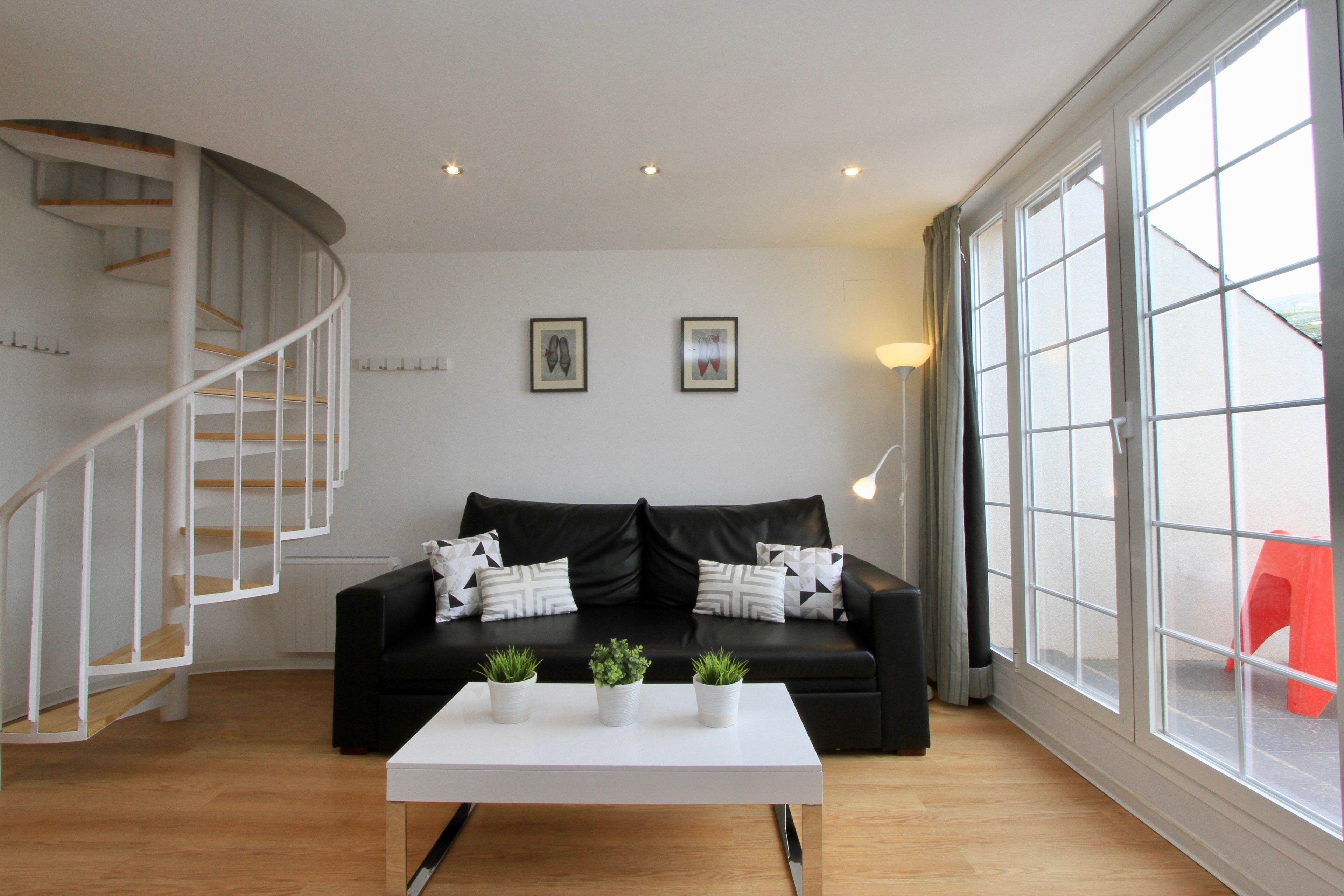 A modern living room with a black leather sofa, white coffee table, and potted plants. There is a spiral staircase on the left and large windows on the right.
