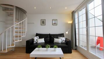 A modern living room with a black leather sofa, white coffee table, and potted plants. There is a spiral staircase on the left and large windows on the right.