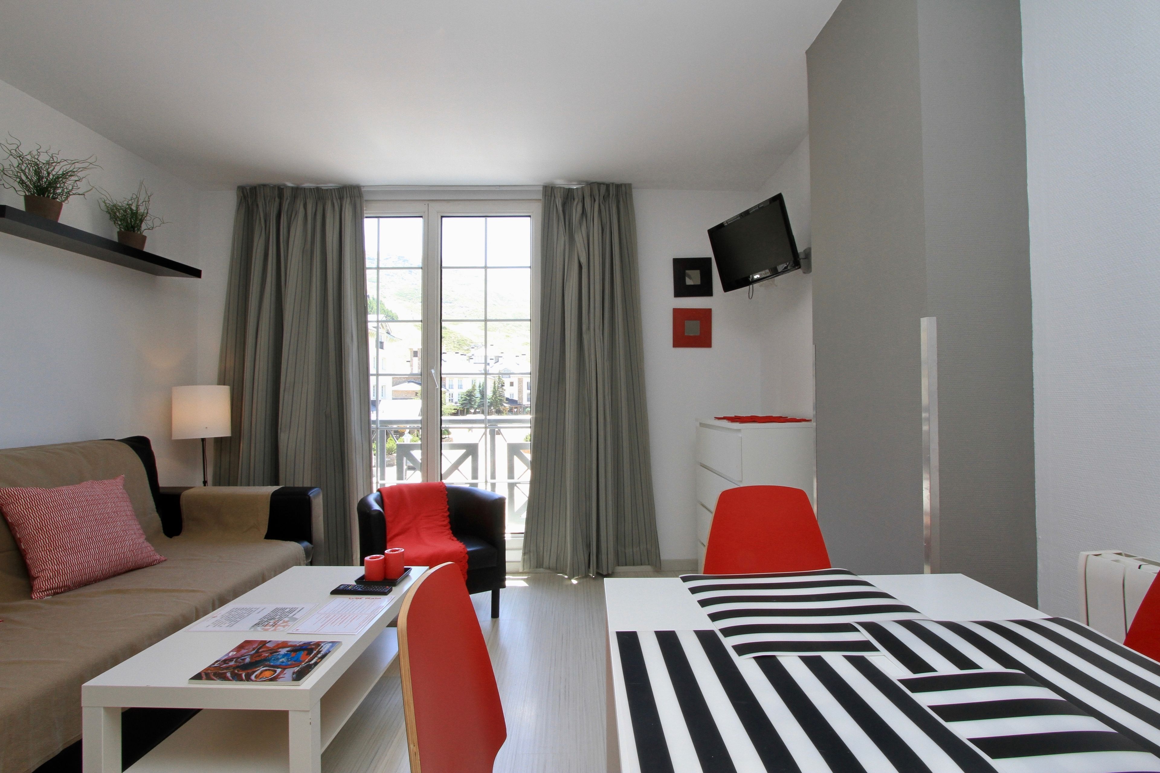 A modern hotel room with a sofa, a dining table, two red chairs, a bed with a striped bedspread, a TV mounted on the wall, and a window with curtains.
