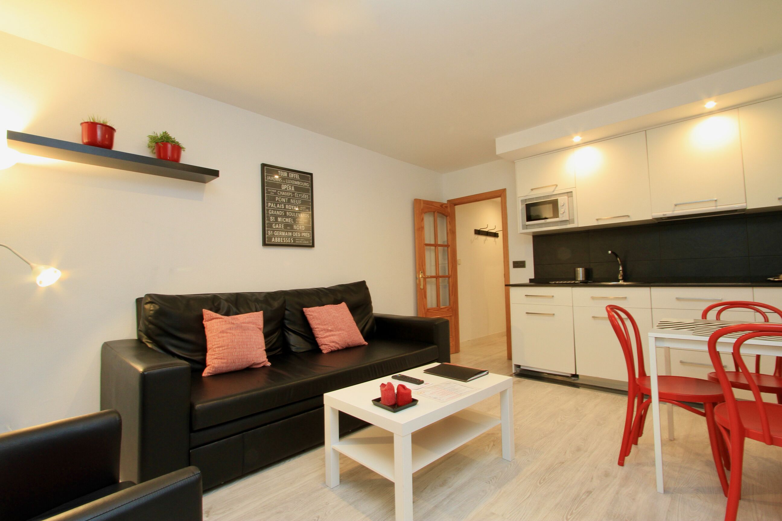 Een moderne woonkamer met een zwarte leren bank, een witte salontafel en rode stoelen. Er is een keuken met witte kasten en een magnetron.