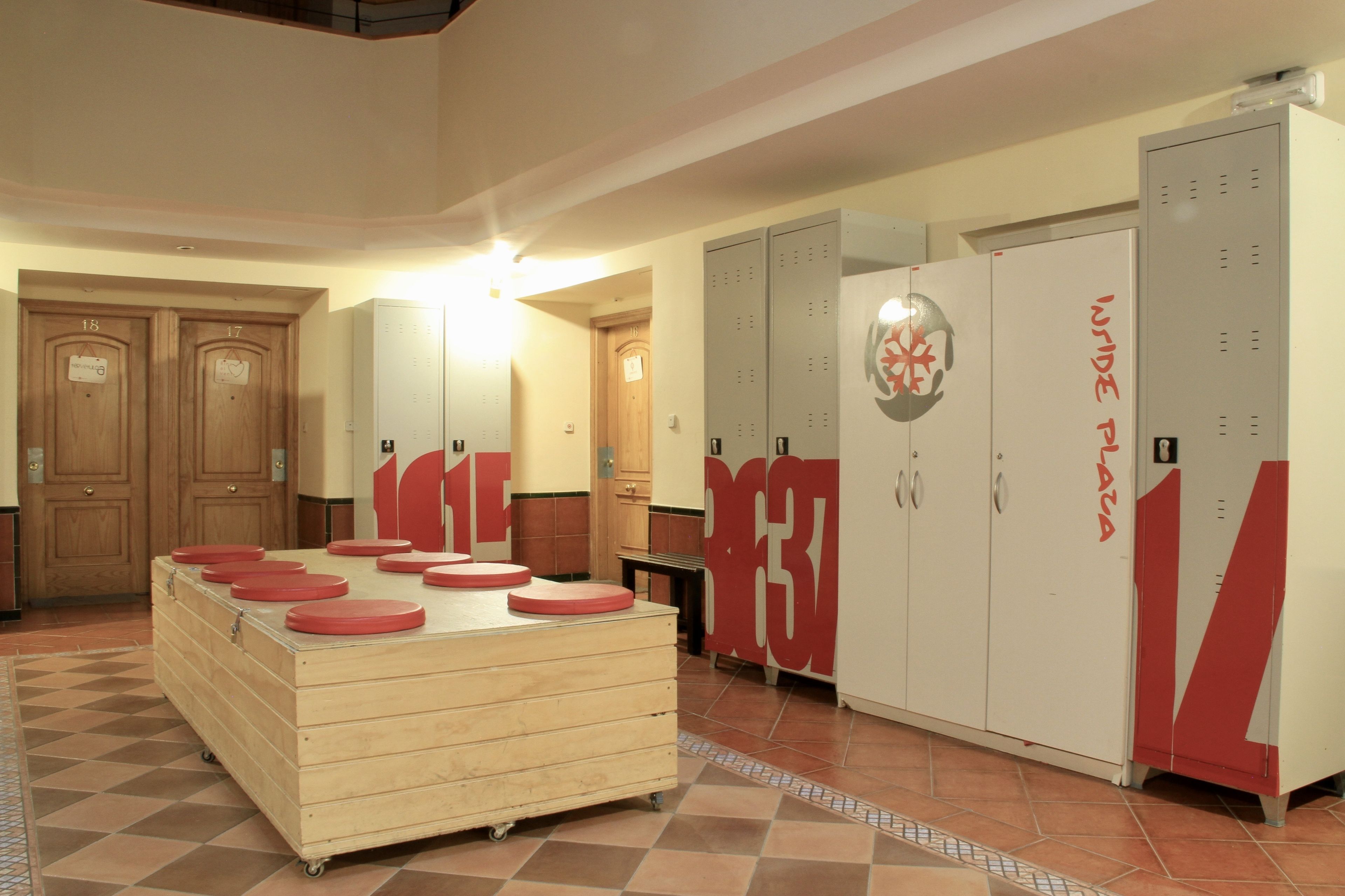 A hotel lobby with wooden lockers, a central wooden table with red stools, and a tiled floor.