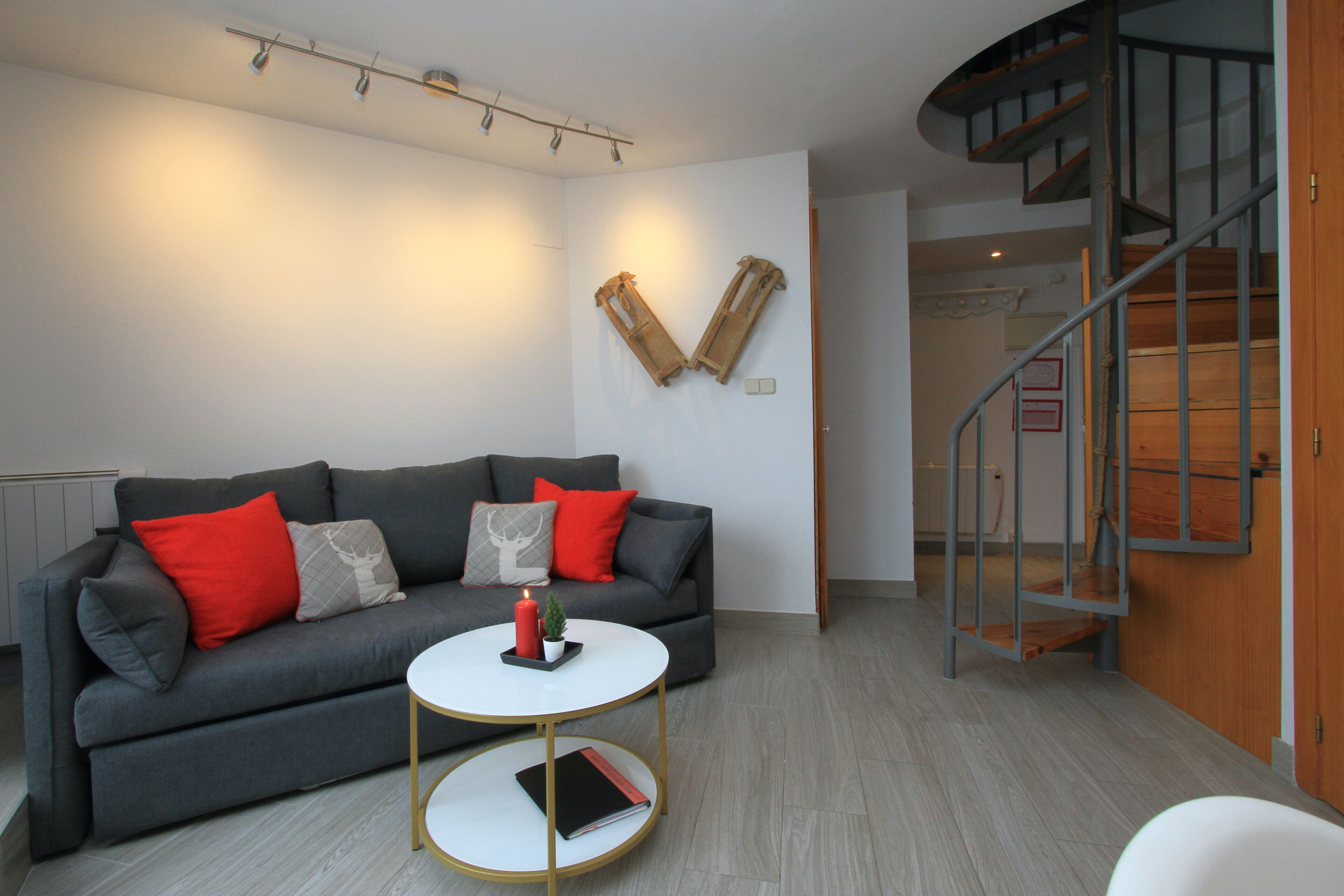 Un salón moderno con un sofá gris, cojines rojos y grises, una mesa de centro blanca y una escalera espiral.