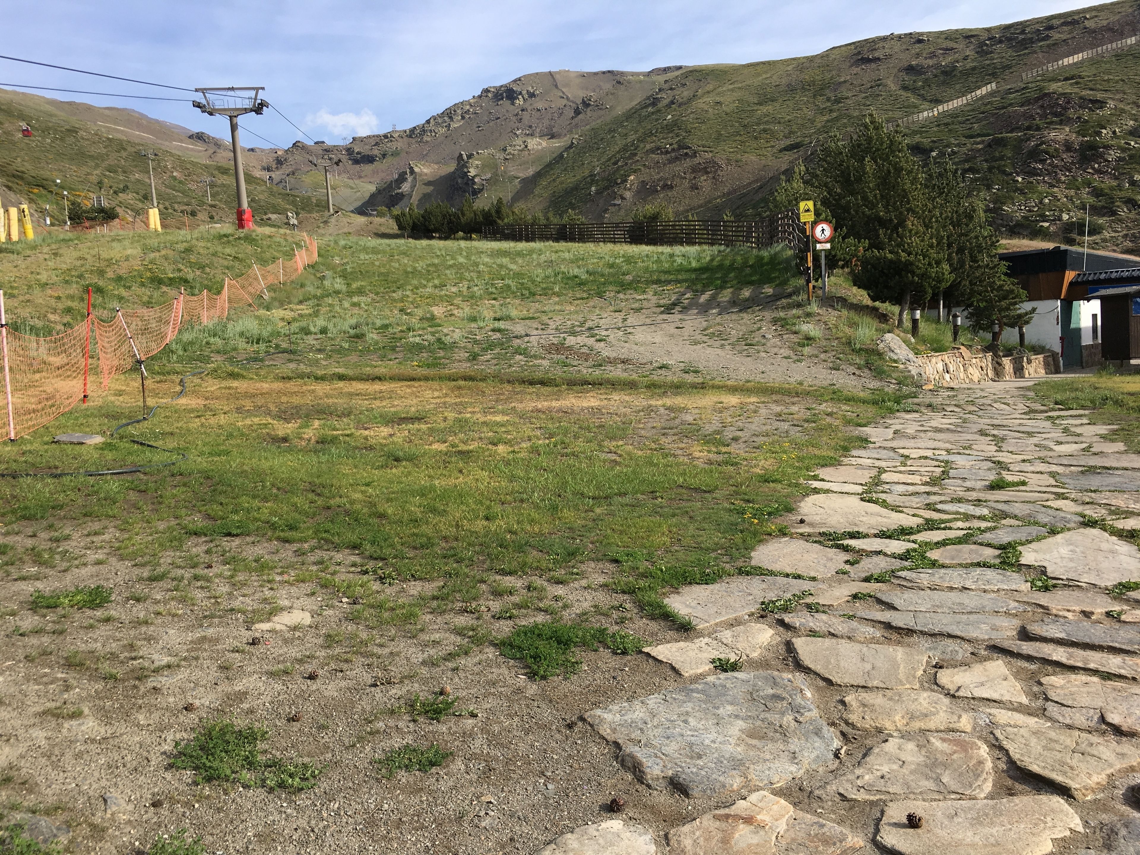 A stone pathway leading uphill with a fenced area and a wooden building in the background.