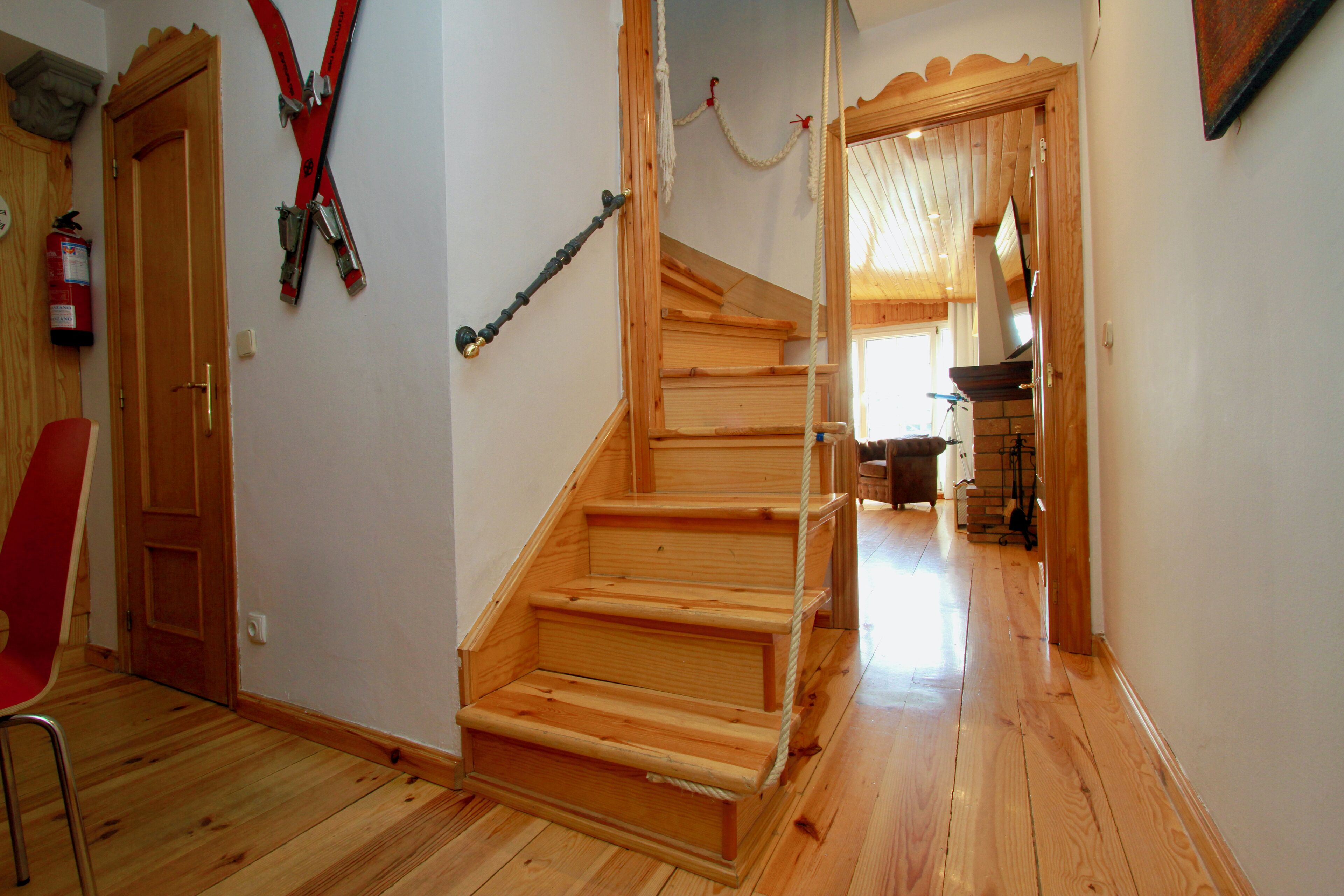 Una escalera de madera con barandilla de cuerda, una puerta de madera y una silla roja en un pasillo con suelo de madera.