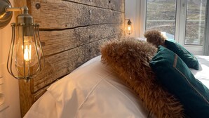 A bedroom with a rustic wooden headboard, two bedside lamps, fluffy pillows, and a view of a mountainous landscape through a large window. - Inside Plaza Sierra Nevada (Monachil)