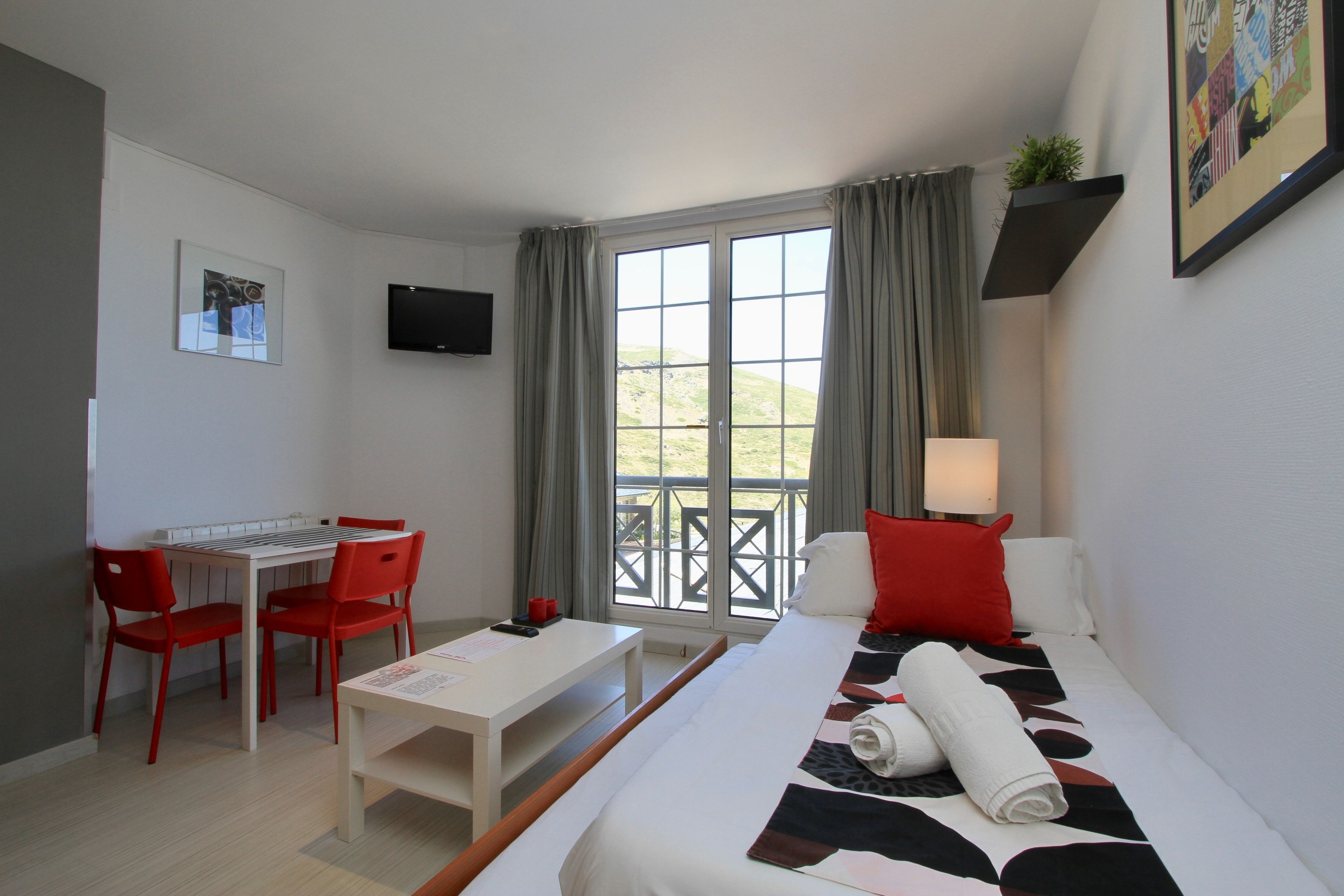 A hotel room with a bed, a desk, two red chairs, a white table, a TV mounted on the wall, and a window with curtains.