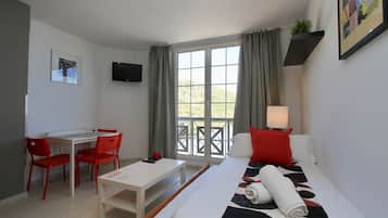 A hotel room with a bed, a desk, two red chairs, a white table, a TV mounted on the wall, and a window with curtains.