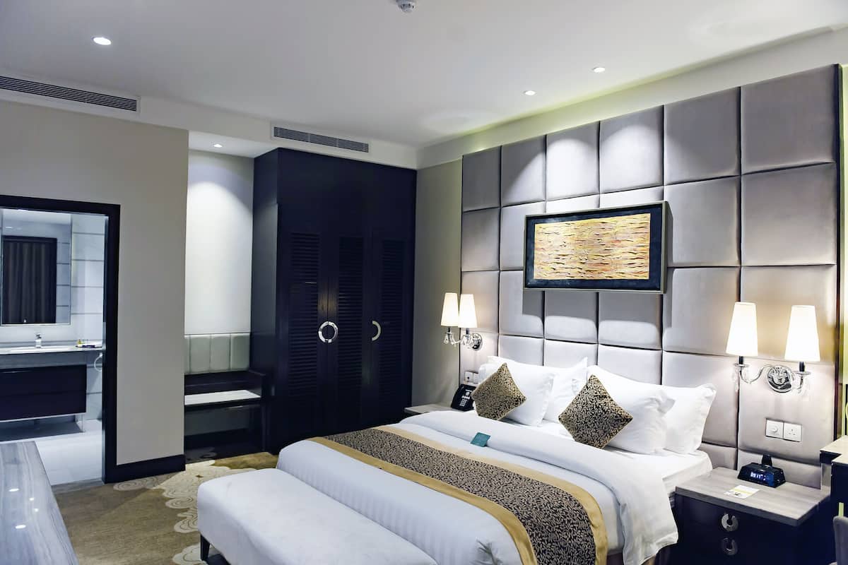 A modern hotel room with a large bed, bedside lamps, a headboard with a mounted artwork, a built-in wardrobe, and a separate bathroom area.