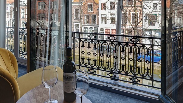 A table with two wine glasses and a bottle of wine, a yellow chair, and a view of buildings through large windows. - Hotel Royal Bridges (Delft)