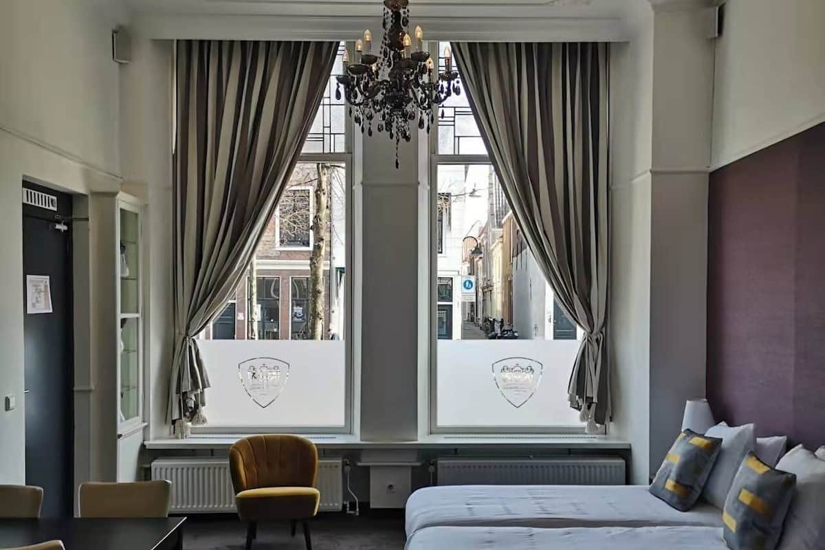 A hotel room with two beds, a chandelier, a dining table with chairs, and large windows with curtains.