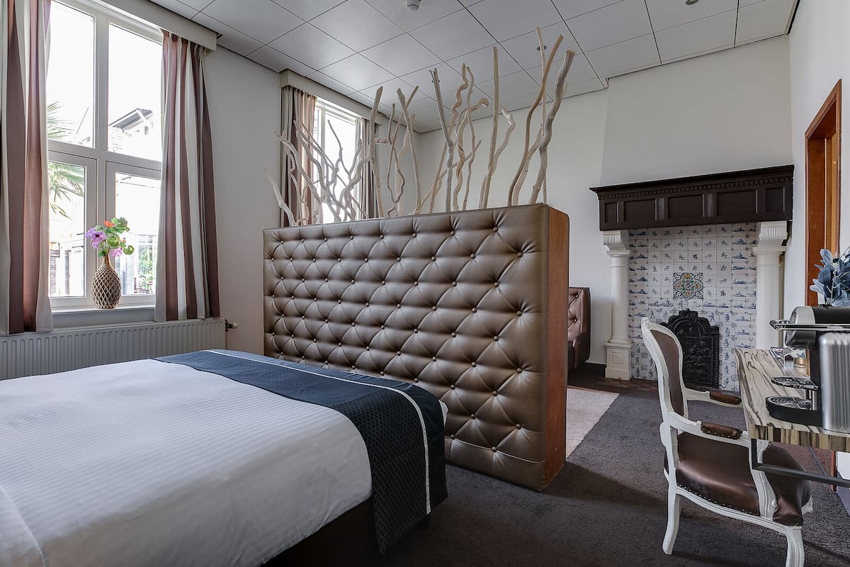 A hotel room with a large tufted headboard, a bed with a white and blue bedspread, a wooden side table, a chair, and a fireplace with patterned tiles.