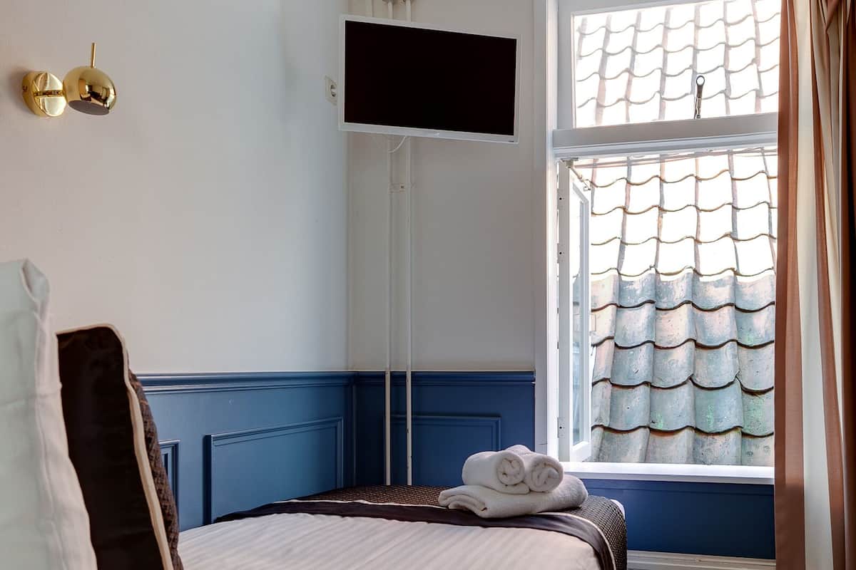 A neatly made bed with a white and brown duvet, a flat-screen TV mounted on the wall, and a window with curtains.