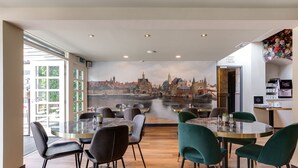 A restaurant with round tables and chairs, a mural of a cityscape, and a view of a river. - Hotel Royal Bridges (Delft)